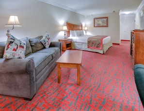 Habitación de hotel con un sofá gris, una mesa de centro de madera, una cama con colcha estampada en rojo y verde, y una mesita de noche de madera con lámparas.
