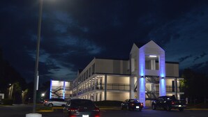 Un immeuble à plusieurs étages, doté d’une tour centrale imposante, de nombreux balcons et d’un espace de stationnement où sont garées des voitures.