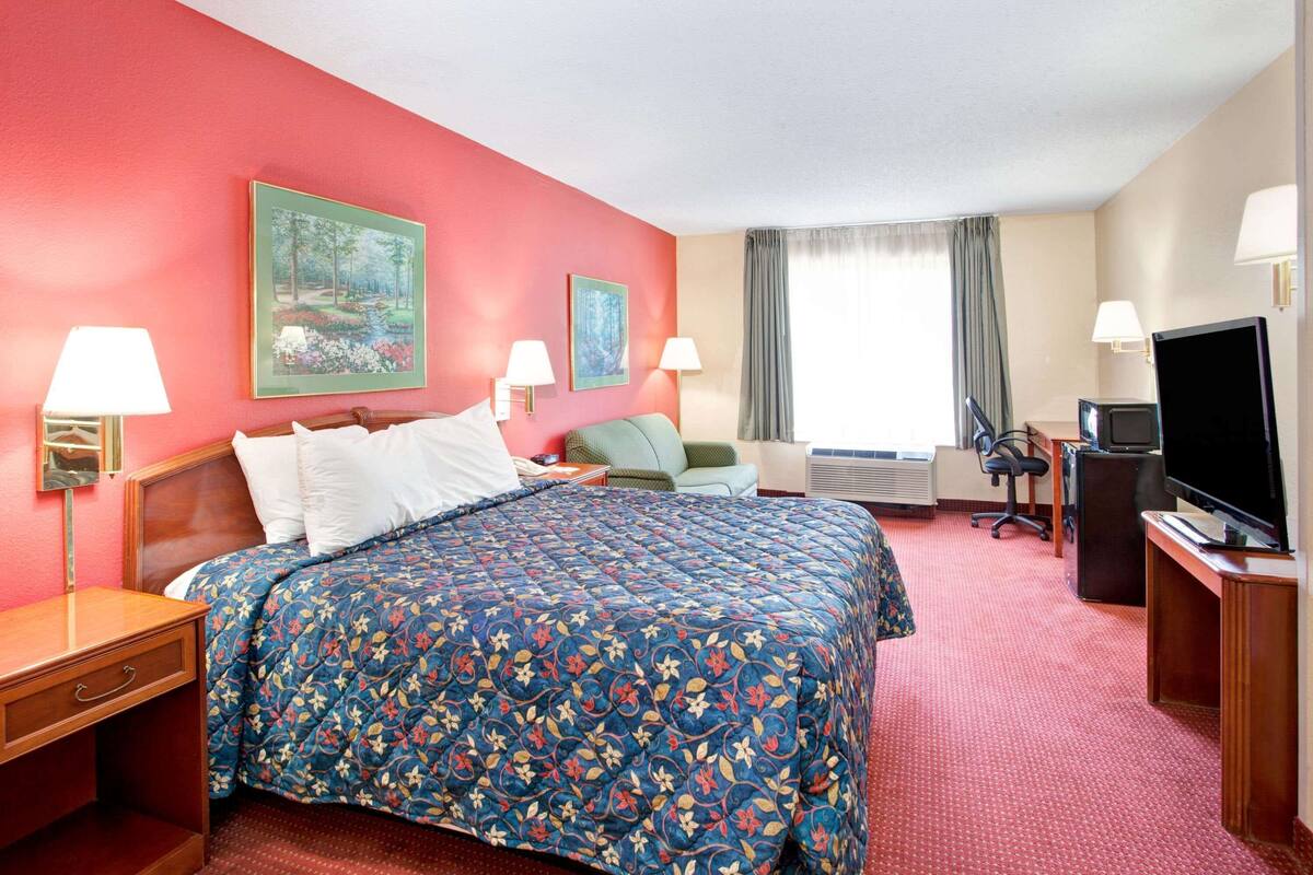 Une chambre d’hôtel avec un grand lit, deux tables de chevet avec des lampes, une télévision, un bureau avec un ordinateur et deux tableaux au mur.