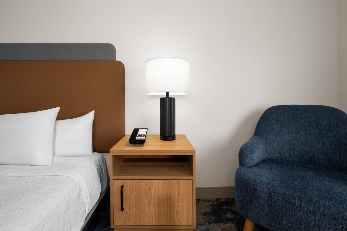 A hotel room with a bed, a nightstand with a lamp, a phone, and a blue chair.