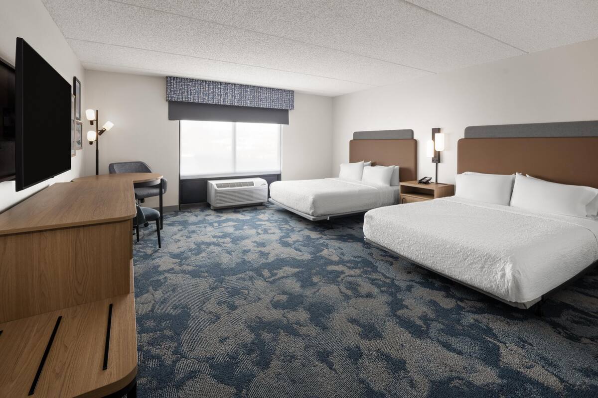 A hotel room with two beds, a desk, a TV, and a window.