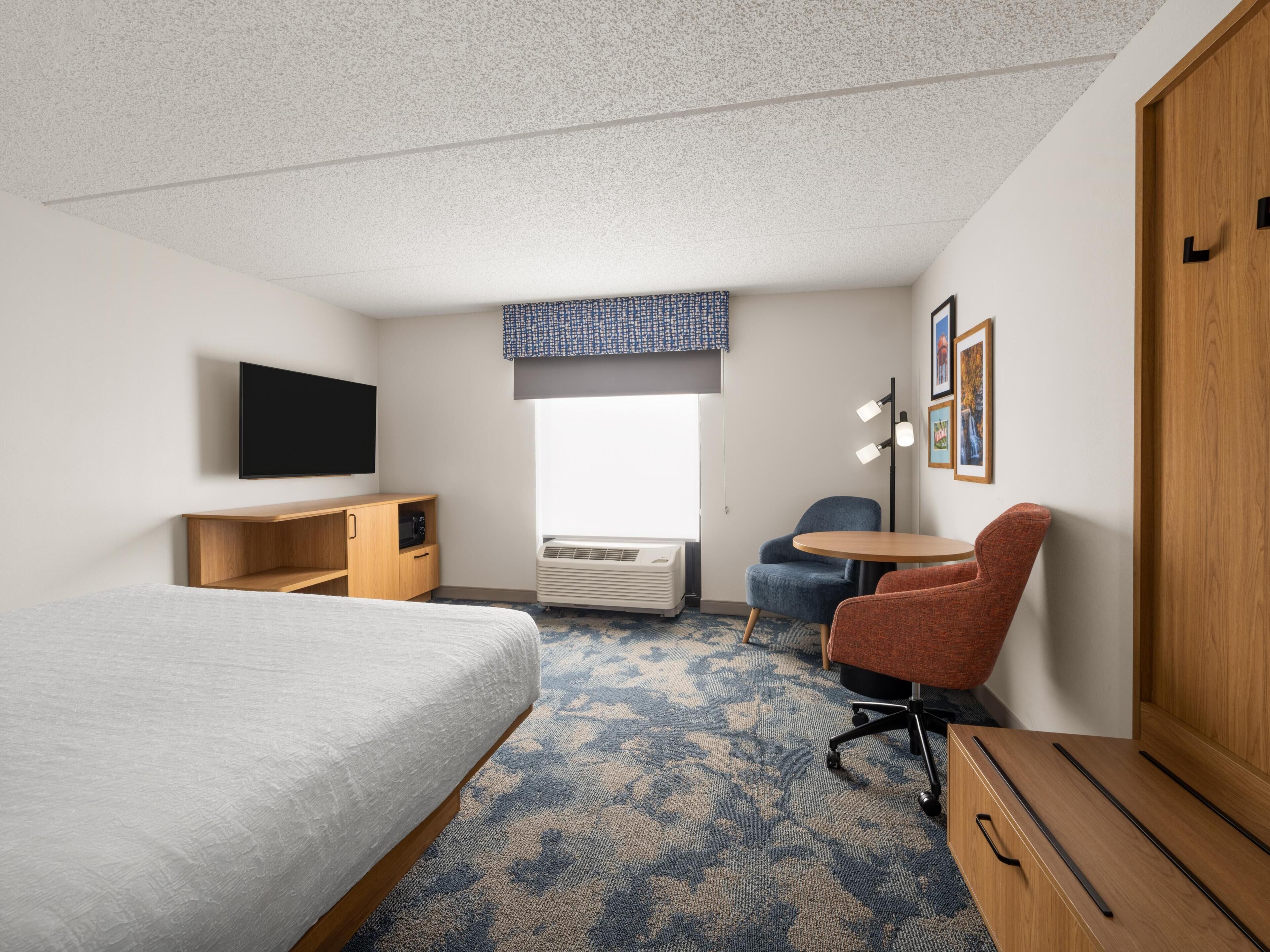 A hotel room with a bed, a desk, a chair, a small table, a TV, an air conditioner, and a wardrobe.