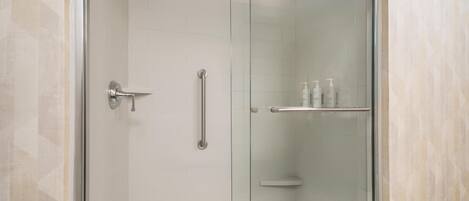 A glass-enclosed shower with a handheld showerhead, a door with a handle, and a shelf with toiletries.