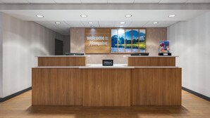 Un mostrador de recepción con un cartel que dice 'Welcome to Hampton', una pantalla digital y una insignia de 'Hilton Honors'.