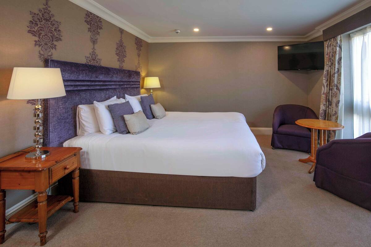 A hotel room with a large bed, a bedside table with a lamp, a chair, and a small round table.