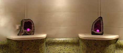 Un baño con paredes y suelo de mosaico, dos lavamanos con encimeras de piedra decorativa y una lámpara en el techo.
