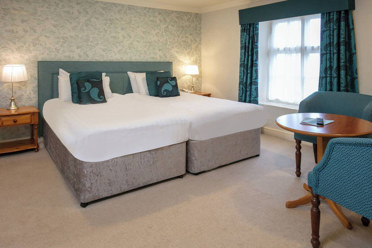 A double bed with two pillows, a bedside table with a lamp, a round wooden table, and a chair.