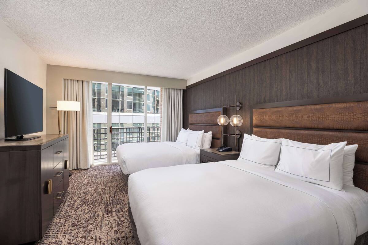 A hotel room with two beds, a TV, a desk, and a window with curtains.