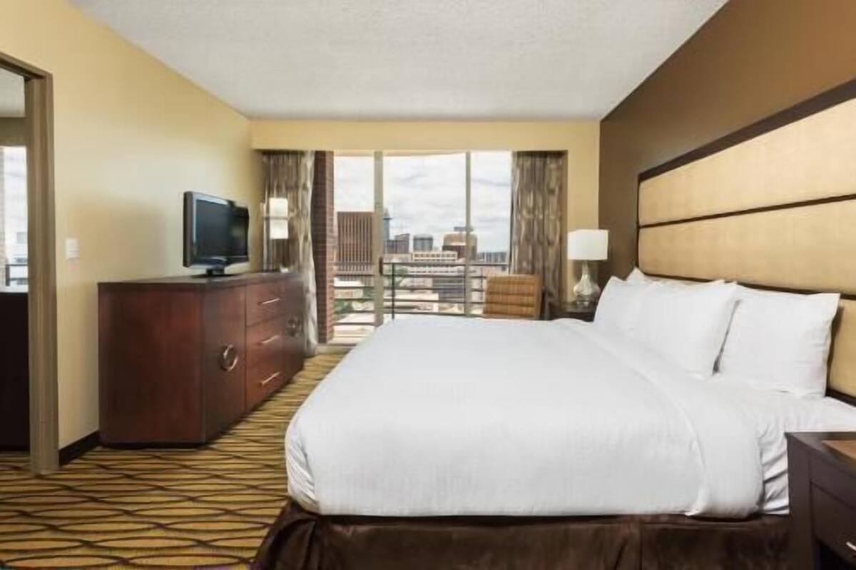 A hotel room with a bed, a TV on a dresser, a view of the city, and a balcony.
