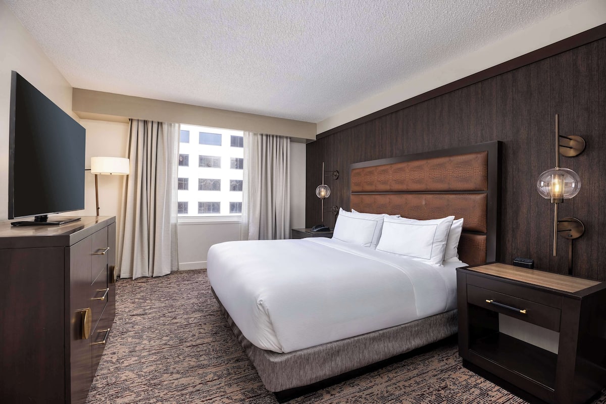 A hotel room with a large bed, a TV, a desk, and a window with curtains.