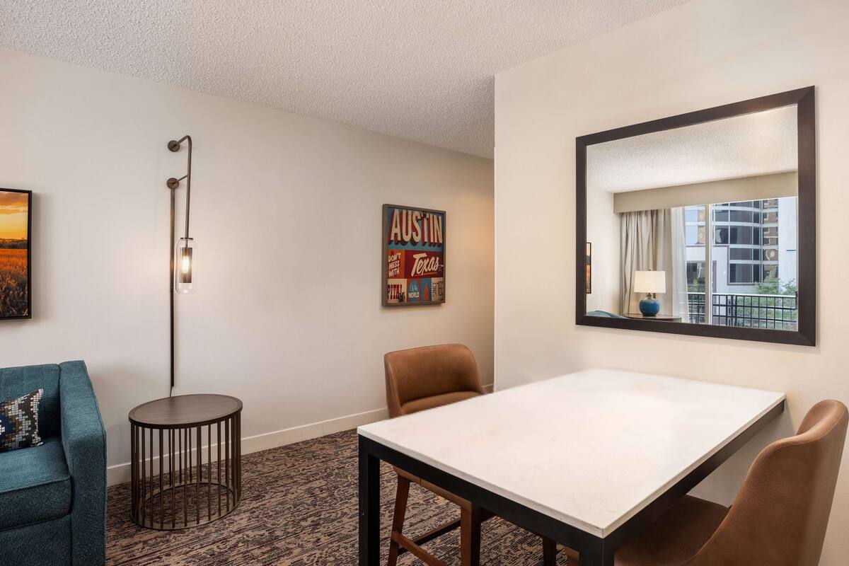 A hotel room with a white table, two brown chairs, a round side table, a floor lamp, a framed sign on the wall, and a window with a view of buildings.