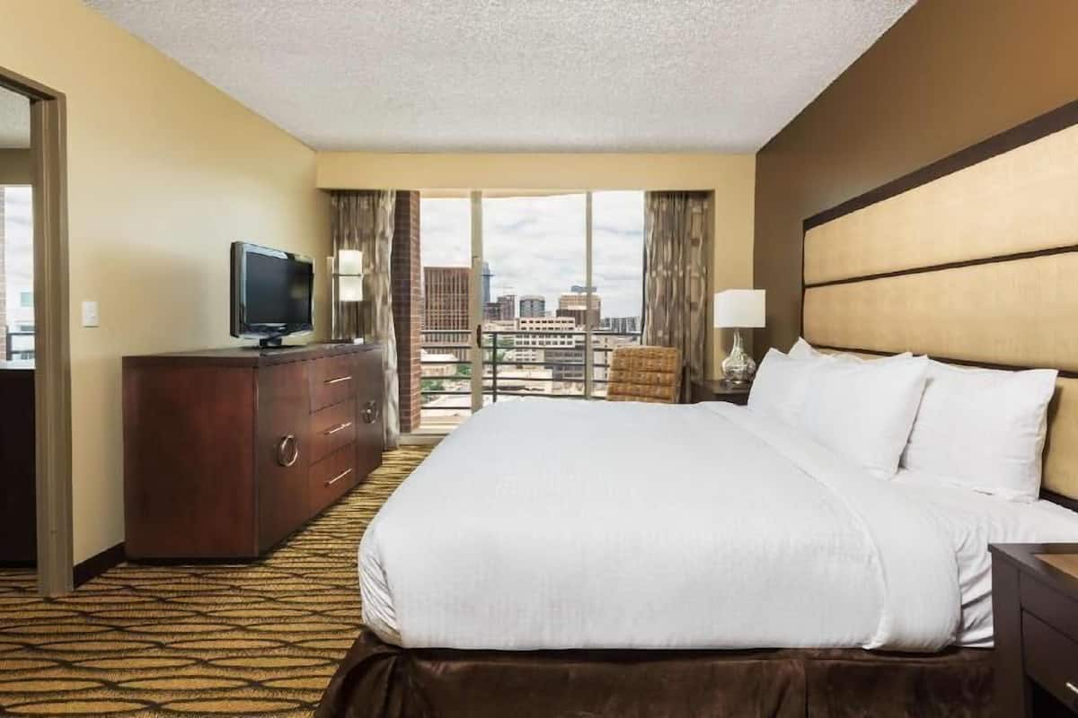 A hotel room with a large bed, a TV on a dresser, a view of the city, and a balcony.