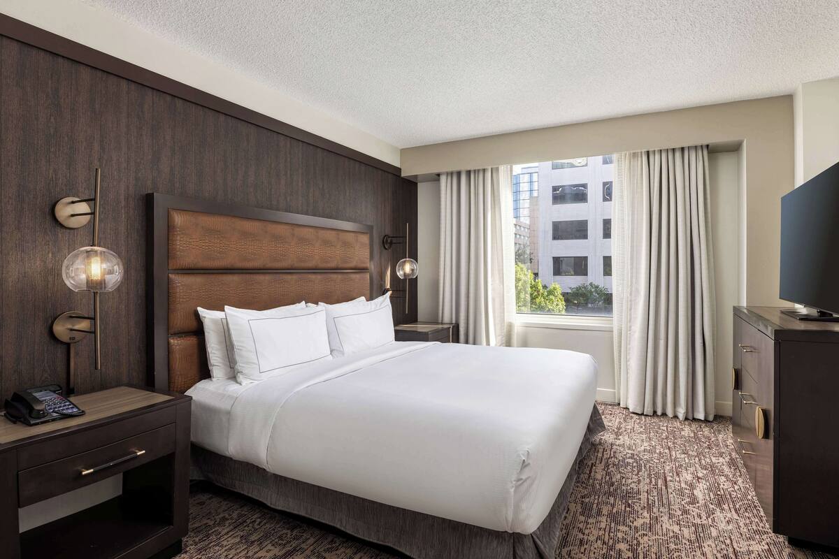 A hotel room with a large bed, a nightstand with a phone, a desk lamp, a TV, and a view of a building through the window.
