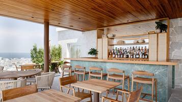 Un área de bar al aire libre con mesas y sillas de madera, una pared de piedra y vistas a la ciudad.