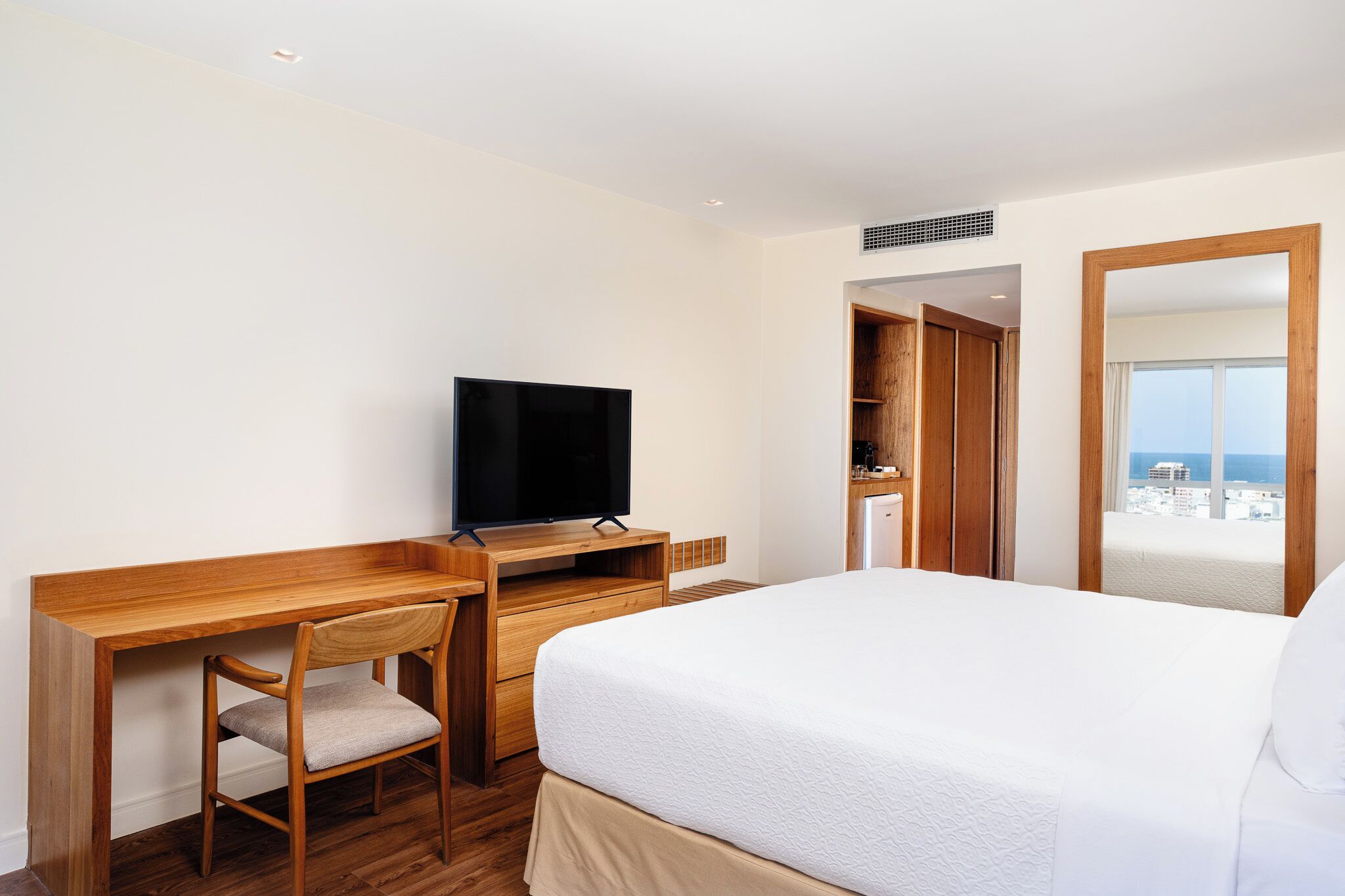 A hotel room with a wooden desk, a television, a bed with white bedding, and a view of the city through a window.