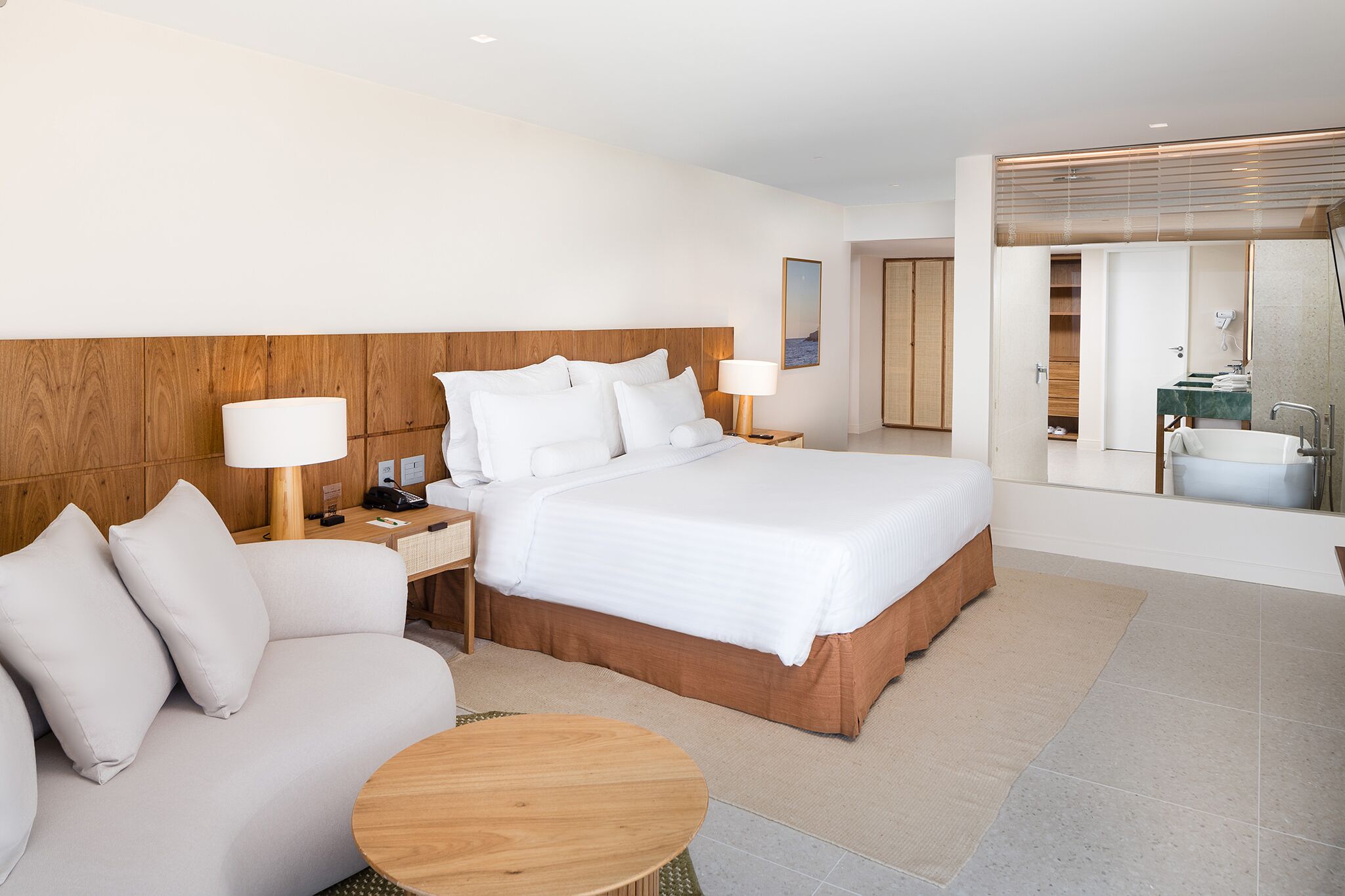 A modern hotel room with a large bed, a sofa, a round wooden table, and a bathroom visible through an open door.