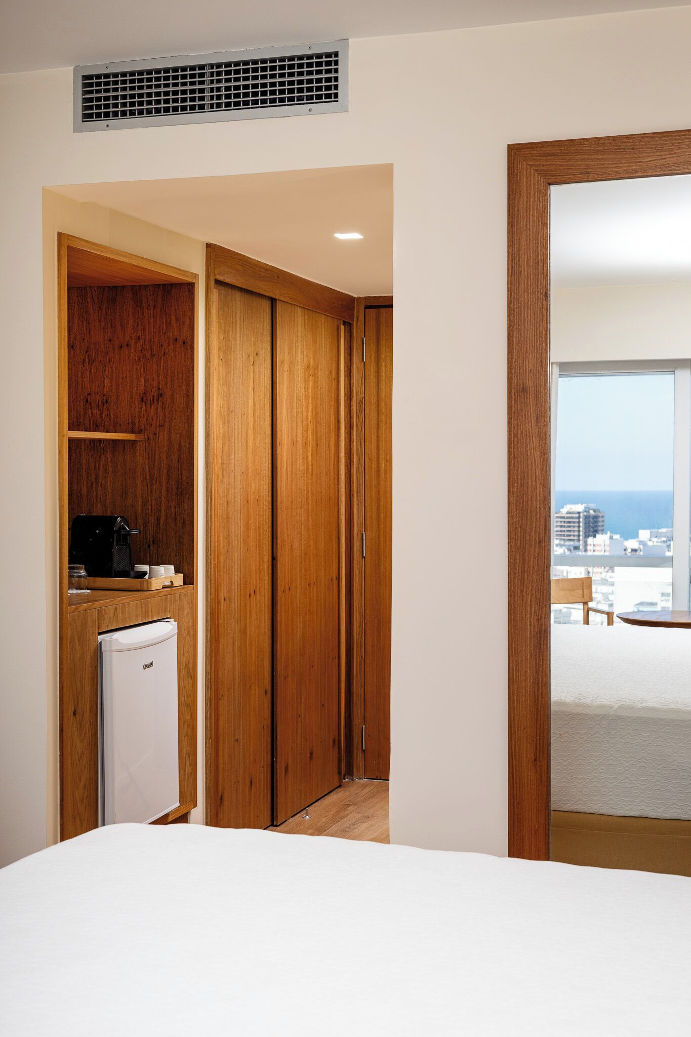 Slaapkamer met een bed, een houten kledingkast, een kleine koelkast en uitzicht op de stad en zee door een groot raam.