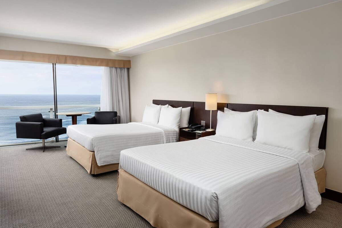 A hotel room with two beds, a desk, a chair, and a view of the ocean.