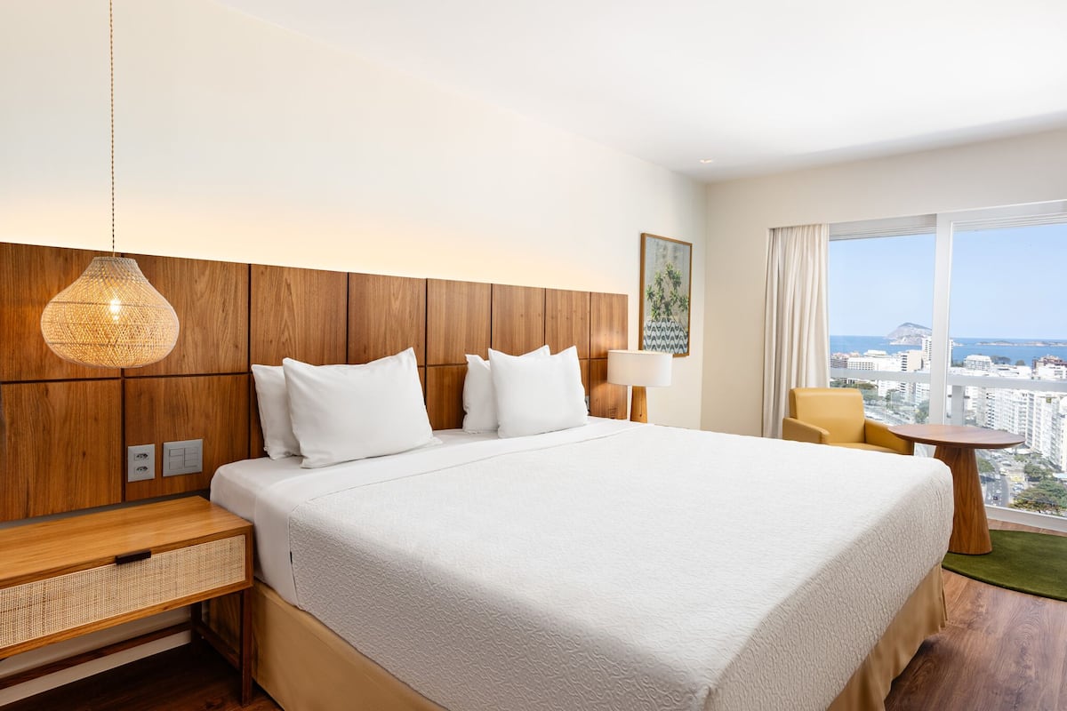 A hotel room with a large bed, wooden headboard, a small table, a chair, and a view of the city through large windows.