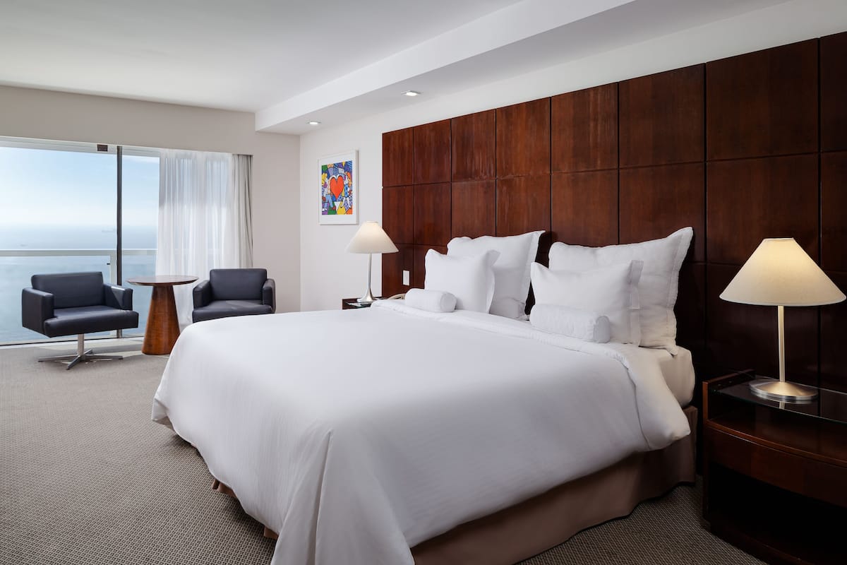 A hotel room with a large bed, two chairs, a small table, a nightstand with a lamp, a large window with curtains, and a colorful artwork on the wall.