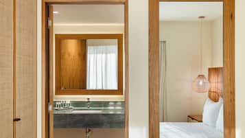 A hotel room with a wooden door, a bathroom with a sink and mirror, a bed with white linens, and a hanging light fixture.