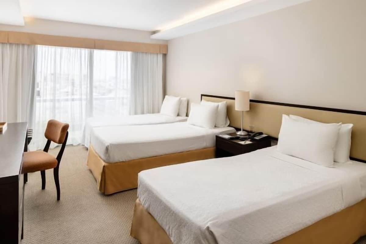 A hotel room with two beds, a desk, a chair, and a nightstand with a lamp.