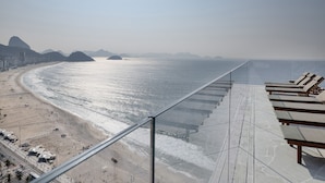 A beachside view from a balcony with sun loungers, a sandy beach, and distant mountains.