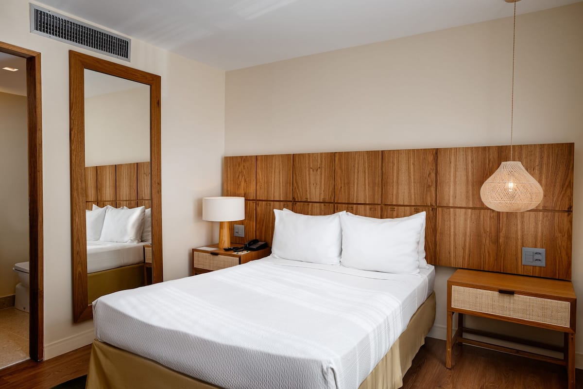 A hotel room with a large bed, wooden headboard, bedside tables with lamps, a mirror, and a woven lamp.