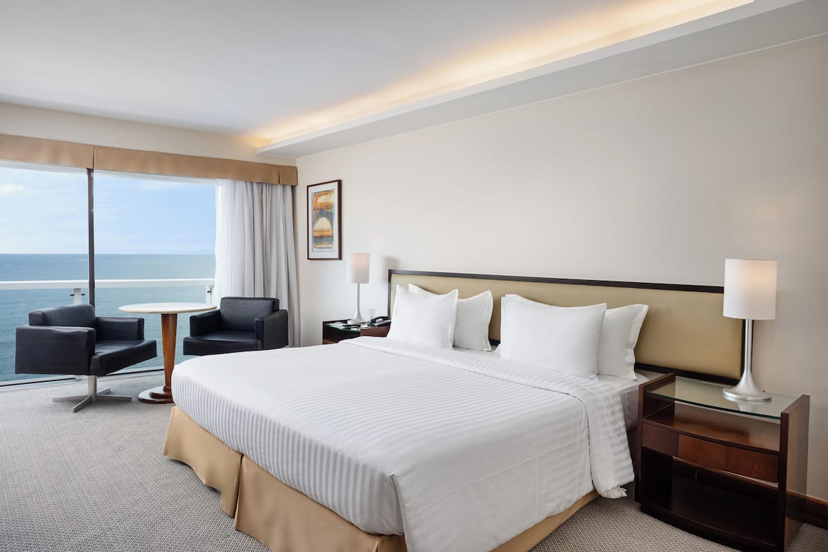A hotel room with a large bed, two bedside tables with lamps, a chair, a small table, and a view of the sea.