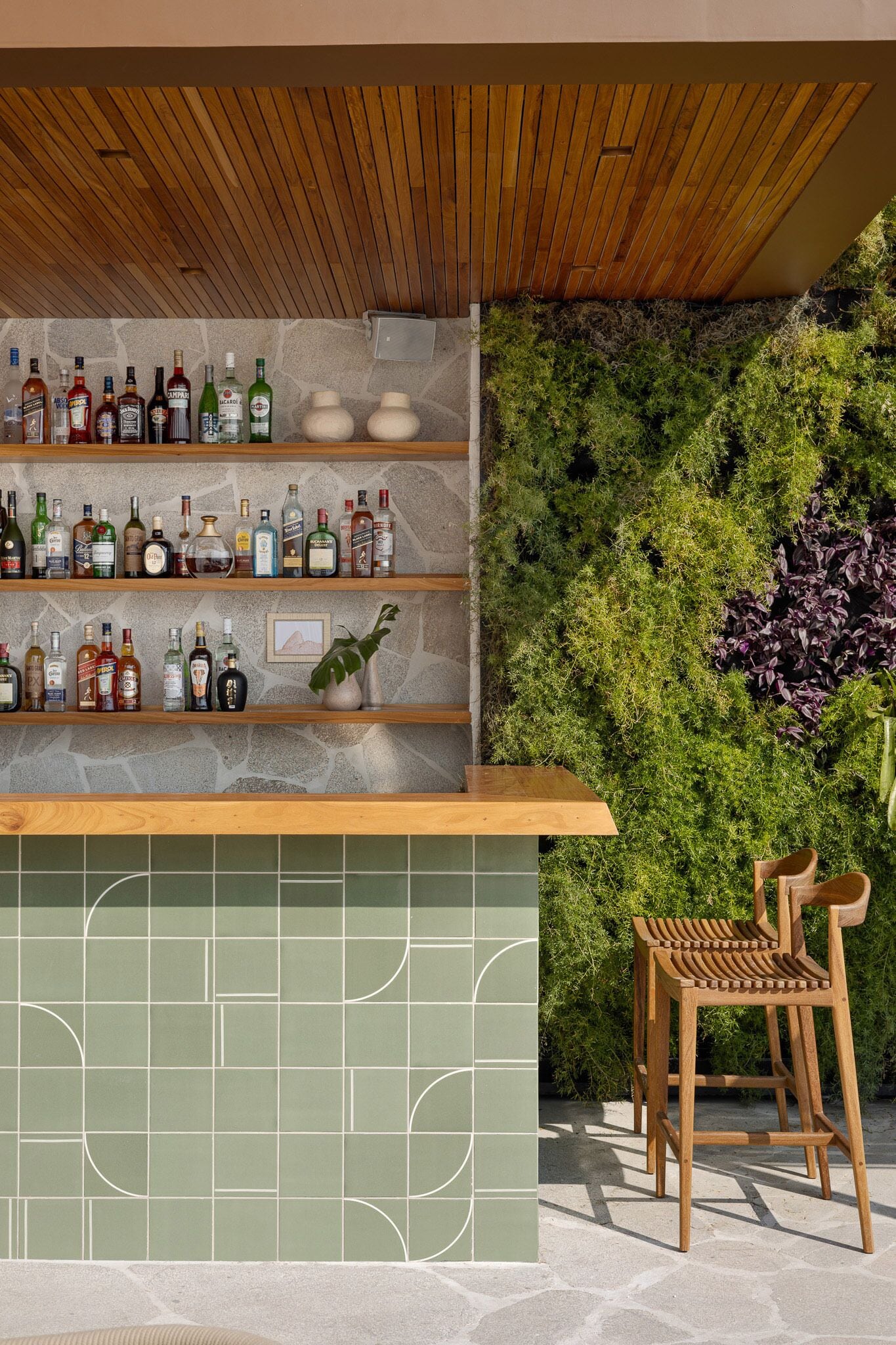 Un espace bar avec des étagères en bois où sont exposées différentes bouteilles, un comptoir recouvert de carreaux verts et une chaise en bois.