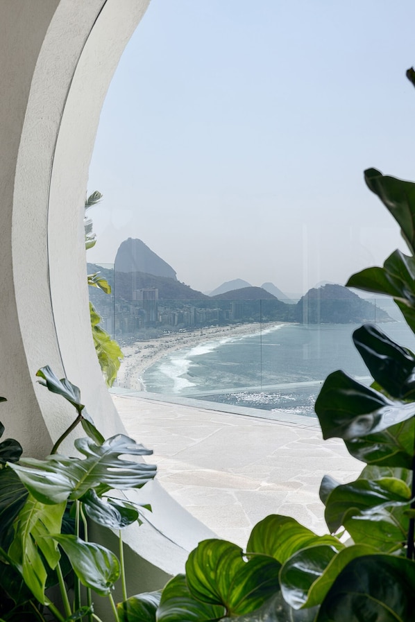 A view of a beach with mountains in the background, framed by a window with green plants in the foreground. - Othon Palace Copacabana Rio (Rio de Janeiro)