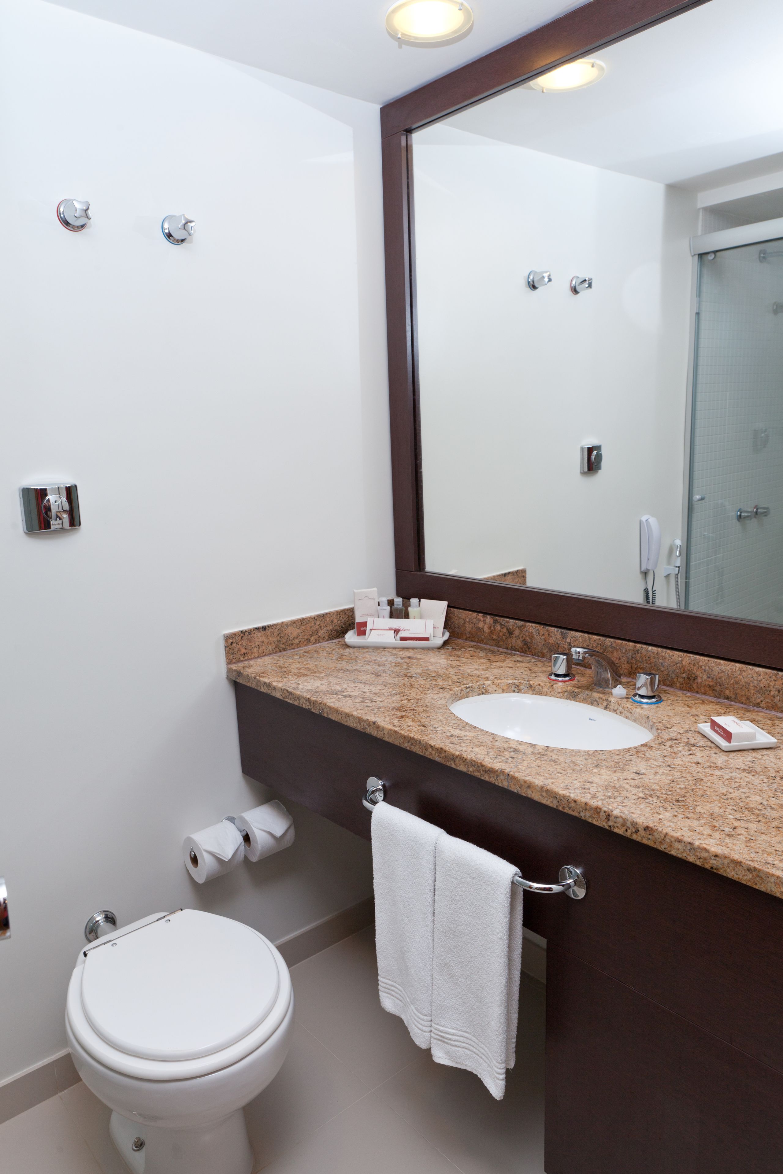 Une salle de bain moderne avec des toilettes blanches, un lavabo doté d’un plan de travail en granit, un grand miroir et un porte-serviettes.