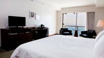 Habitación de hotel con una cama grande, un escritorio con televisor de pantalla plana, una silla, una mesita pequeña y vistas al mar a través de ventanas amplias.