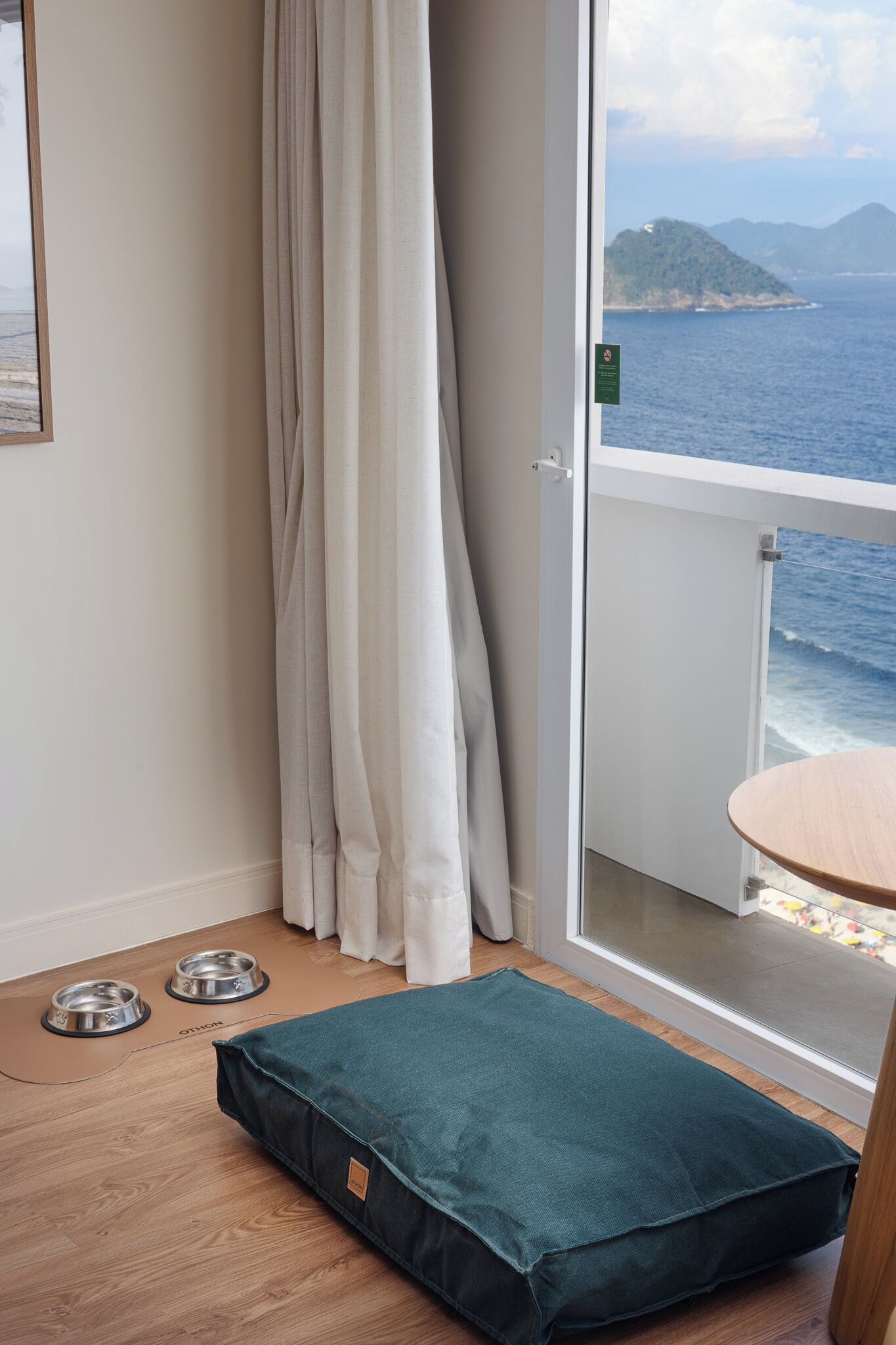 Quarto com piso de madeira, cama para animais de estimação em tom de verde-água, duas tigelas para animais e uma porta de vidro deslizante que dá para uma varanda com vista para o mar e montanhas.