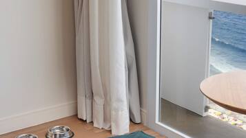Quarto com piso de madeira, cama para animais de estimação em tom de verde-água, duas tigelas para animais e uma porta de vidro deslizante que dá para uma varanda com vista para o mar e montanhas.