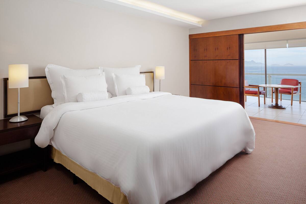 A hotel room with a large bed, two bedside lamps, a wooden wardrobe, and a balcony with a view.