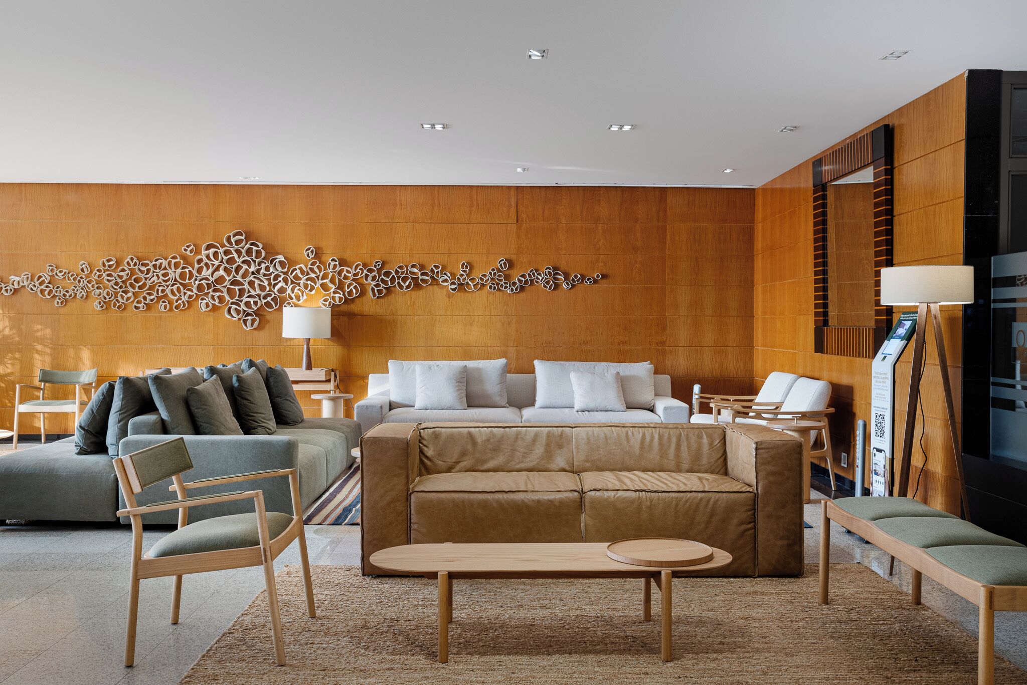 A modern hotel lobby with a wooden wall, a large abstract art piece, a beige sofa, a green armchair, and a wooden coffee table.