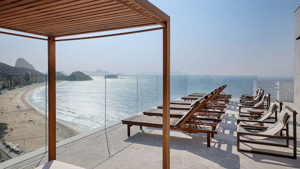 A balcony with a glass railing overlooking a beach and ocean, featuring sun loungers and a pergola.