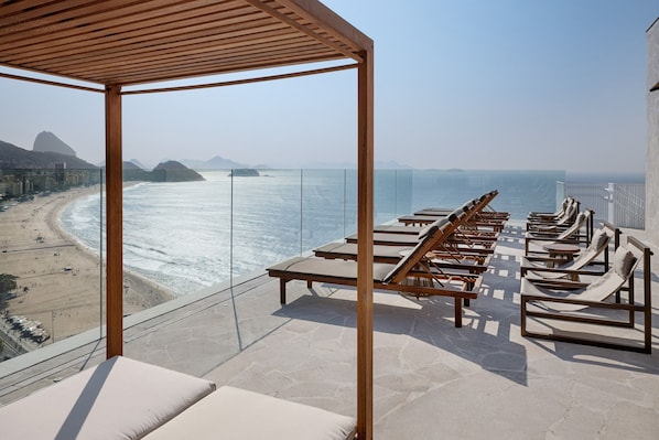 Un balcon doté d’une balustrade en verre, offrant une vue sur une plage et l’océan, et comprenant des chaises longues et une pergola.