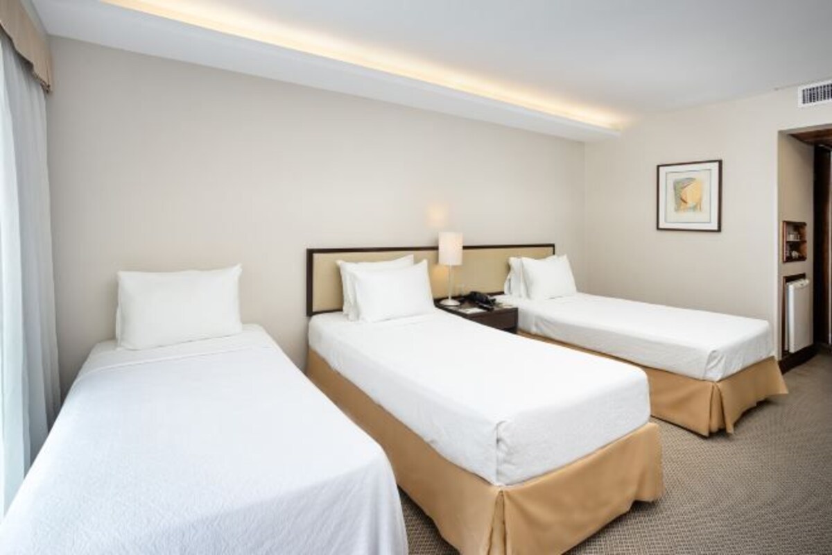 Una habitación de hotel con dos camas individuales, una mesita de noche con lámpara, un cuadro enmarcado en la pared y una puerta que da a otra habitación.