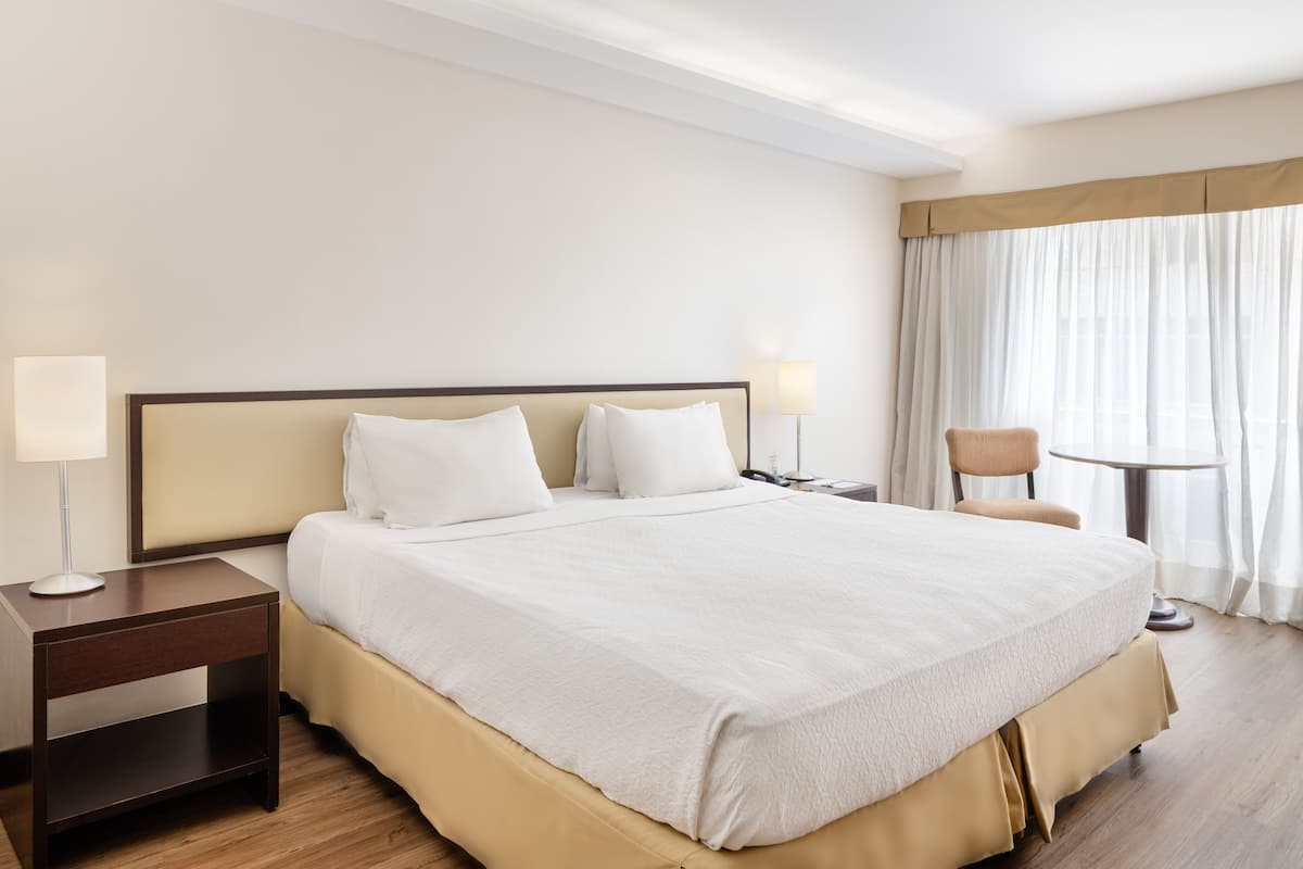 A hotel room with a large bed, a nightstand, a chair, and a window with curtains.