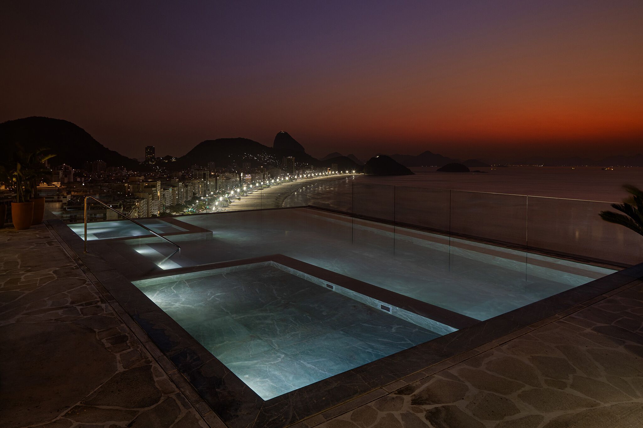 Piscina no terraço com painéis de vidro transparente e vista para a cidade ao entardecer.