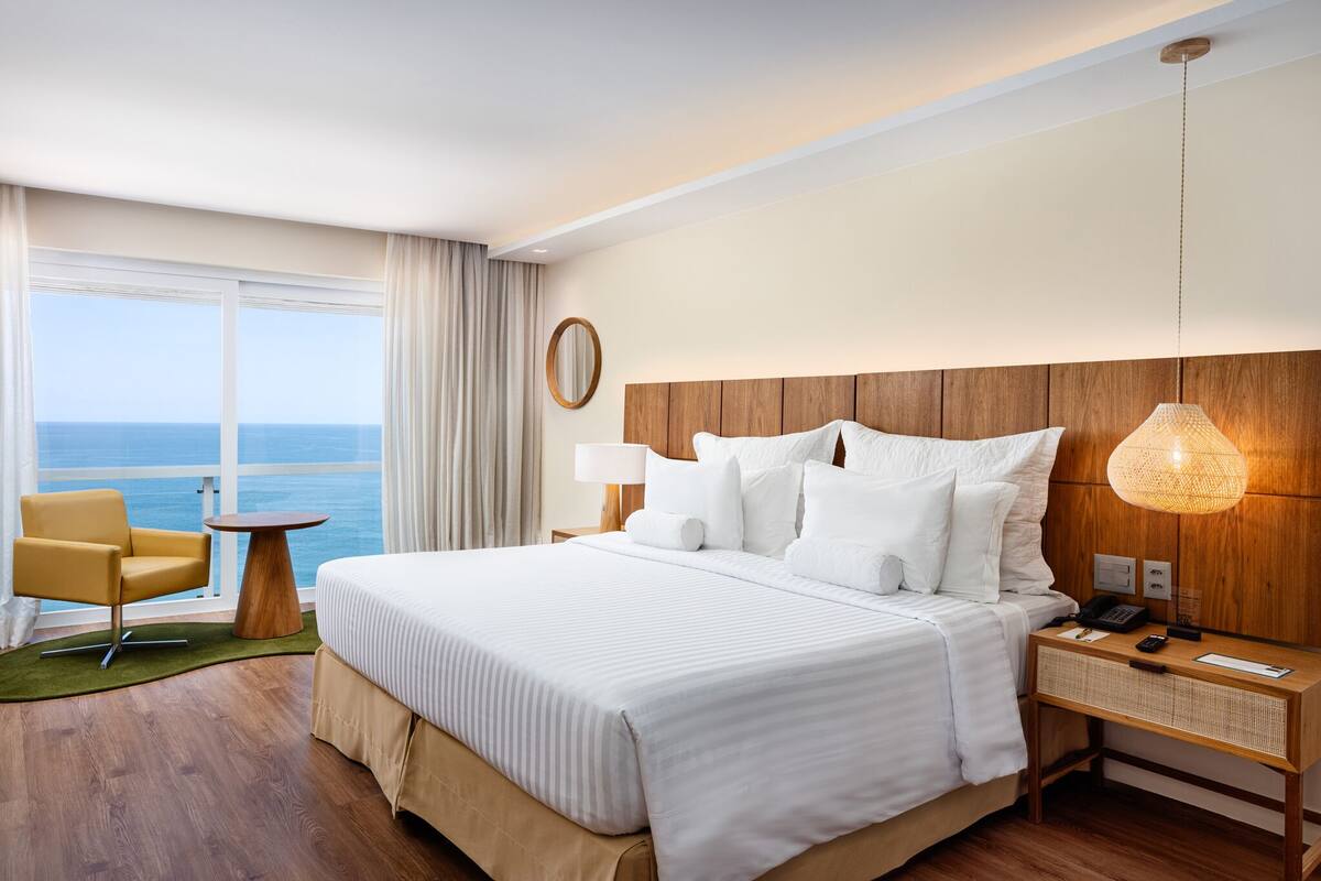 A hotel room with a large bed, a wooden headboard, a bedside table with a lamp, a chair, and a view of the ocean.