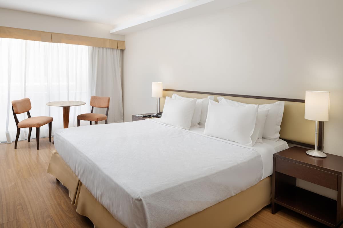 A hotel room with a large bed, two bedside tables, a small round table, two chairs, and a window with curtains.