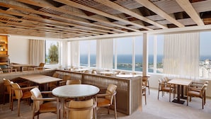 A modern restaurant with high ceilings, wooden beams, and large windows offering a view of the ocean.