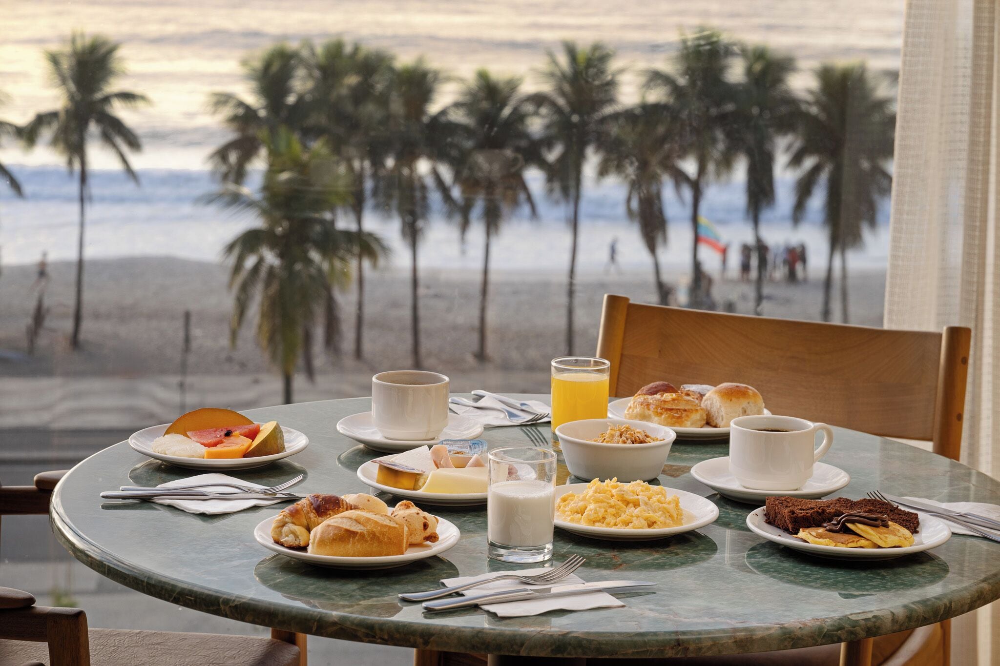 A breakfast table with plates of food, a glass of orange juice, and a cup of coffee, overlooking a beach with palm trees.