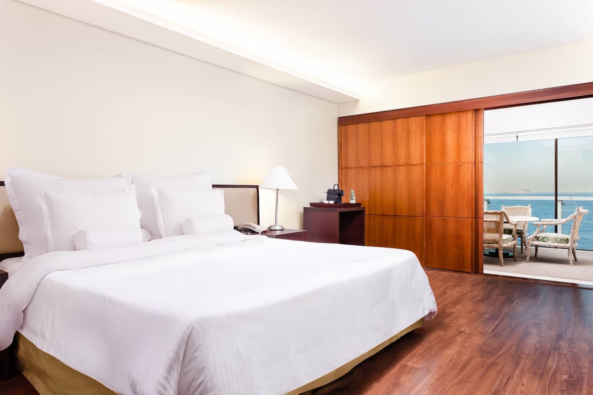 A hotel room with a large bed, a nightstand, a lamp, a wooden wardrobe, and a balcony with a view of the sea.