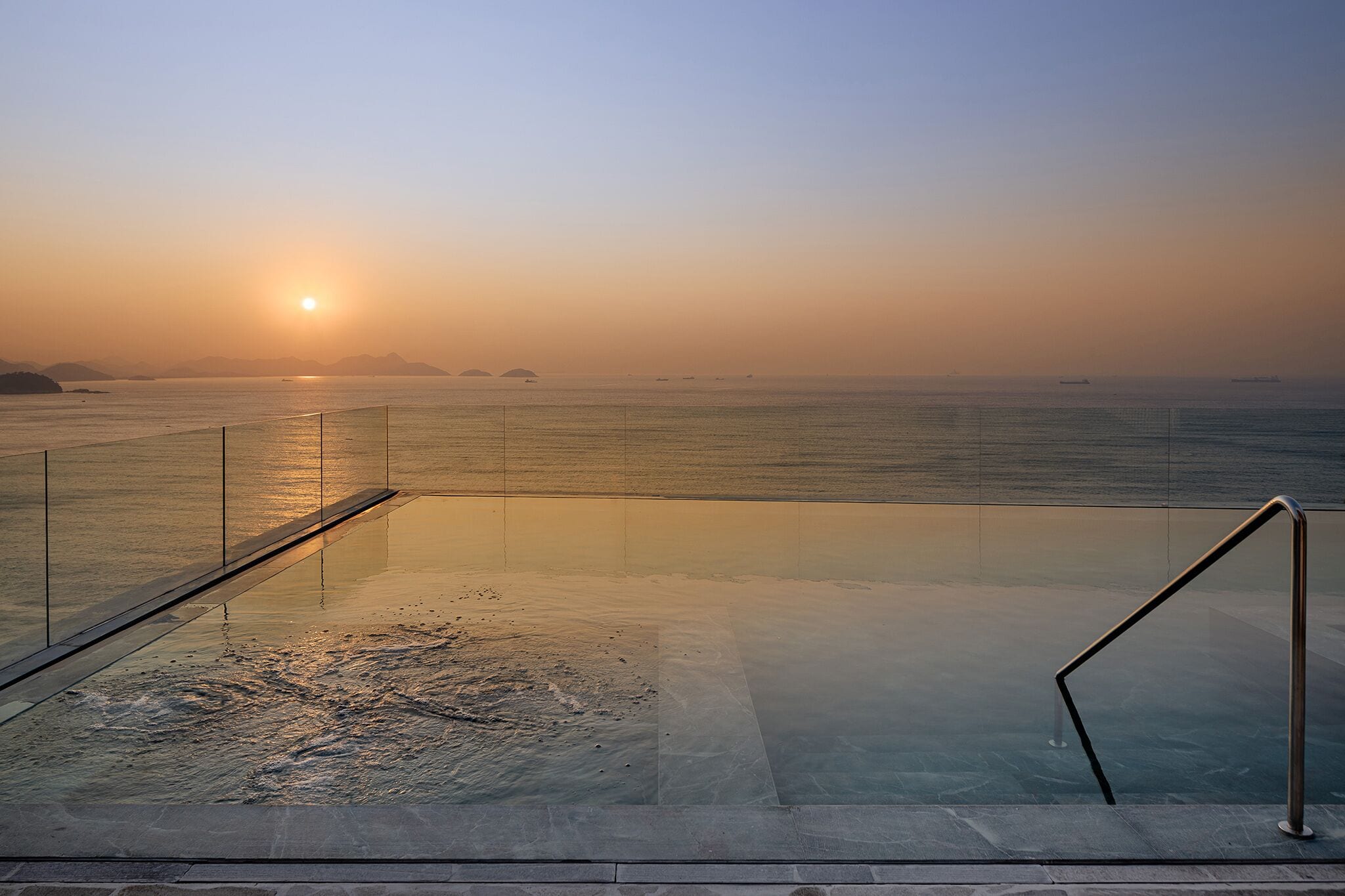 A glass-bottomed infinity pool with a metal railing, overlooking a calm sea and distant mountains during sunset.
