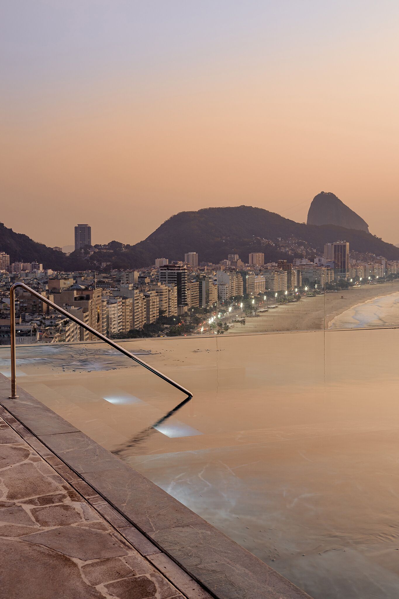 Paisagem costeira urbana com uma montanha imponente ao fundo, uma praia e uma escadaria que desce até a praia.