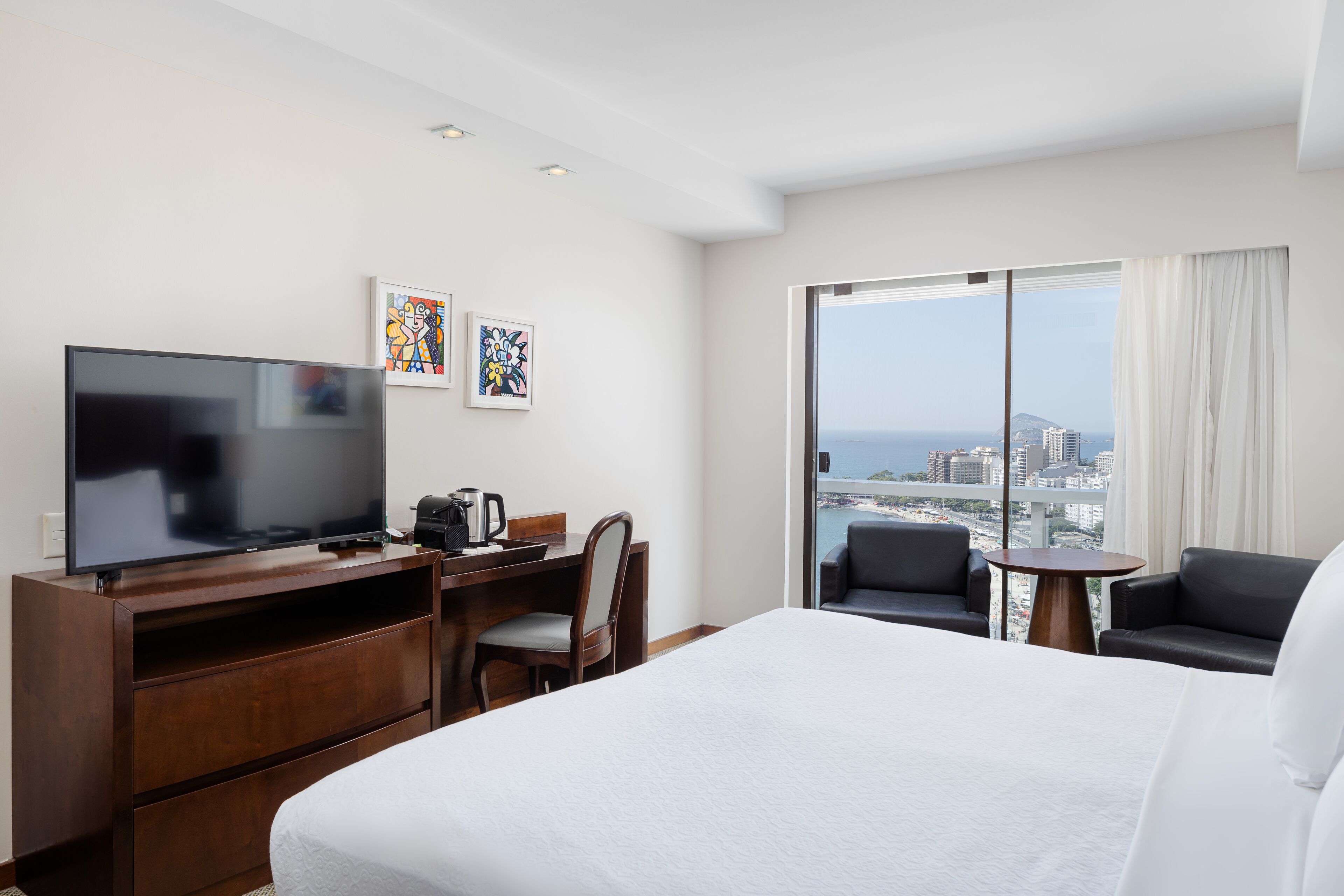 A hotel room with a large bed, a flat-screen TV, a desk with a chair, a coffee maker, and a view of the city and ocean through large windows.